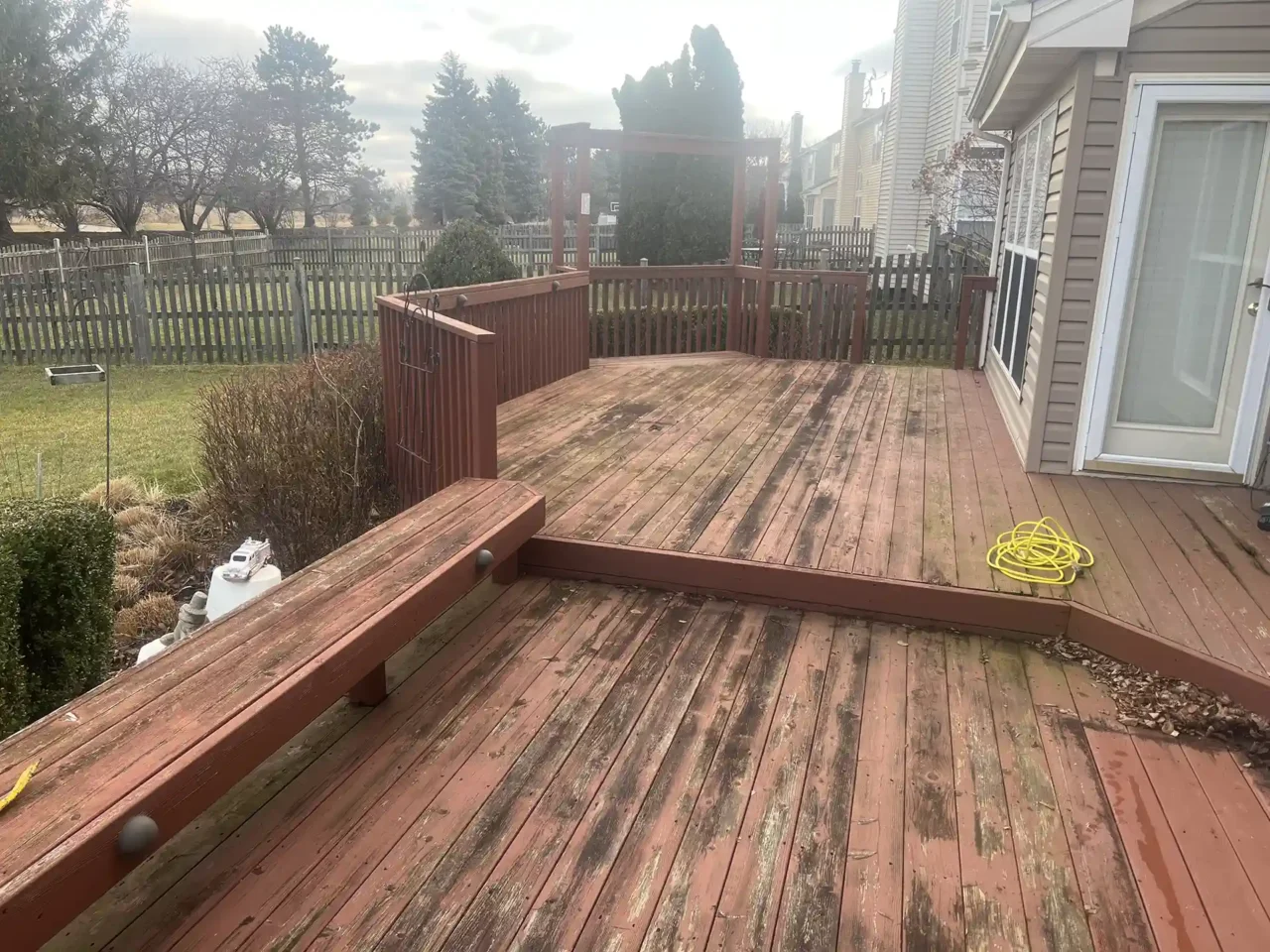 Photo of Weathered red wooden deck in Algonquin before renovation, showing faded boards, built-in bench, and overgrown edges near vegetation.